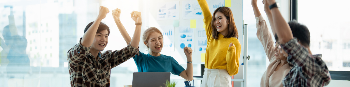 Egy csapat fiatal, lelkes dolgozó ünnepli a sikereket egy irodai környezetben, háttérben diagramokkal és grafikonokkal.