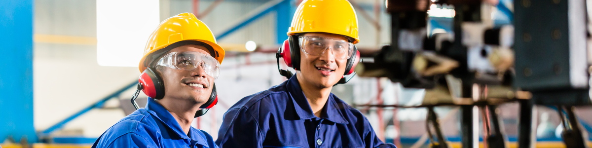 Két mosolygó munkás védőfelszerelésben áll ipari környezetben, gépek között.